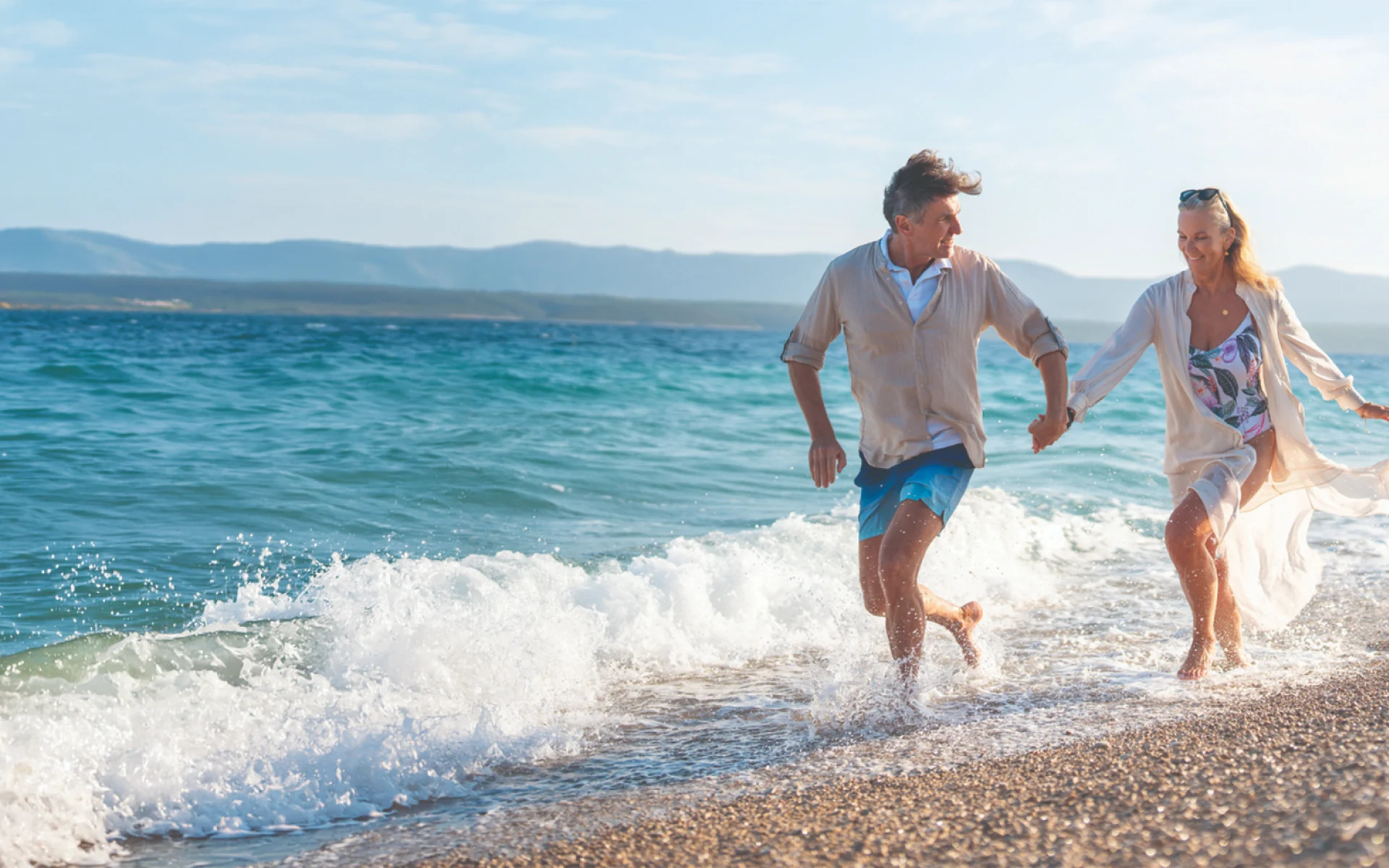 Active older couple running hand-in-hand along a sunlit beach at the water's edge, smiling — outcome-led aspiration
